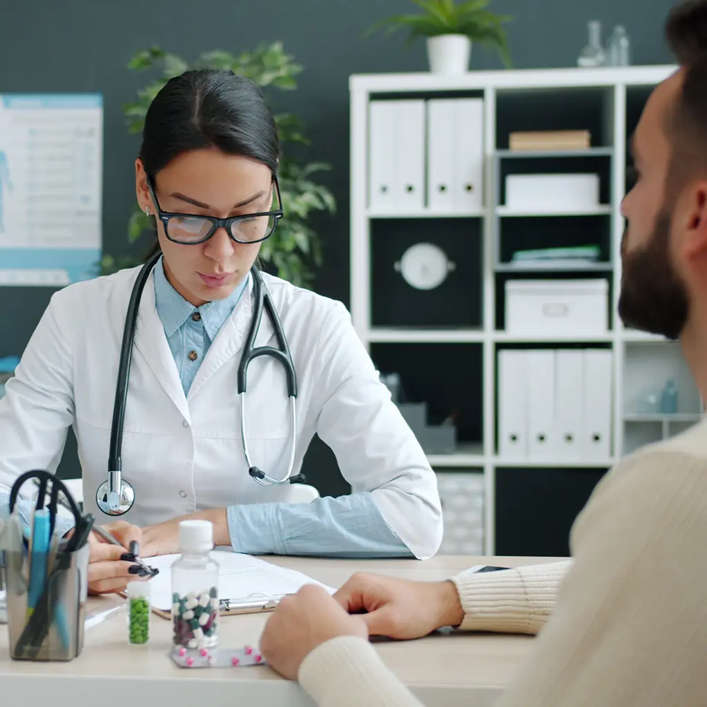Physician taking notes on a clipboard while meeting with a patient at her desk, with medication bottles nearby, during a cost of future care assessment