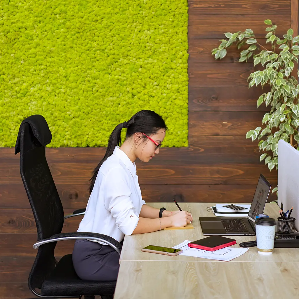 Office employee seated upright in a high-back chair writing notes at her desk workstation with a laptop