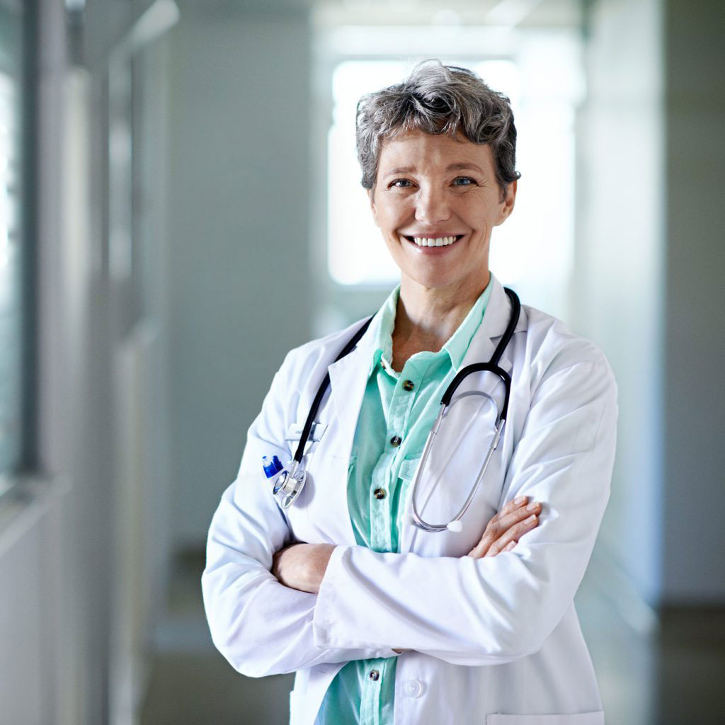 A smiling female medical doctor in a white lab coat with a stethoscope around her neck, standing in a brightly lit clinic hallway.
