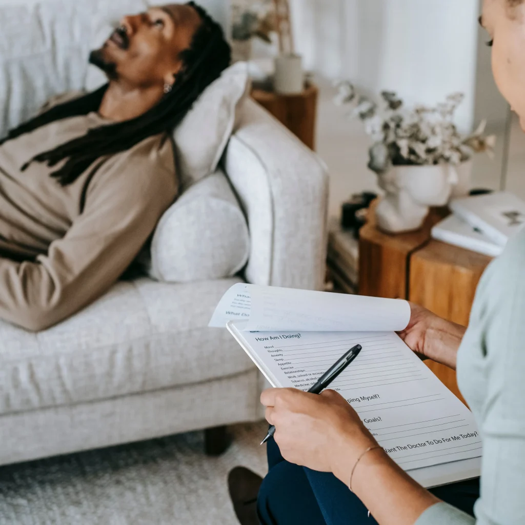 Psychiatrist reviewing a patient assessment form on a clipboard while a client reclines on a couch during an independent psychiatric evaluation