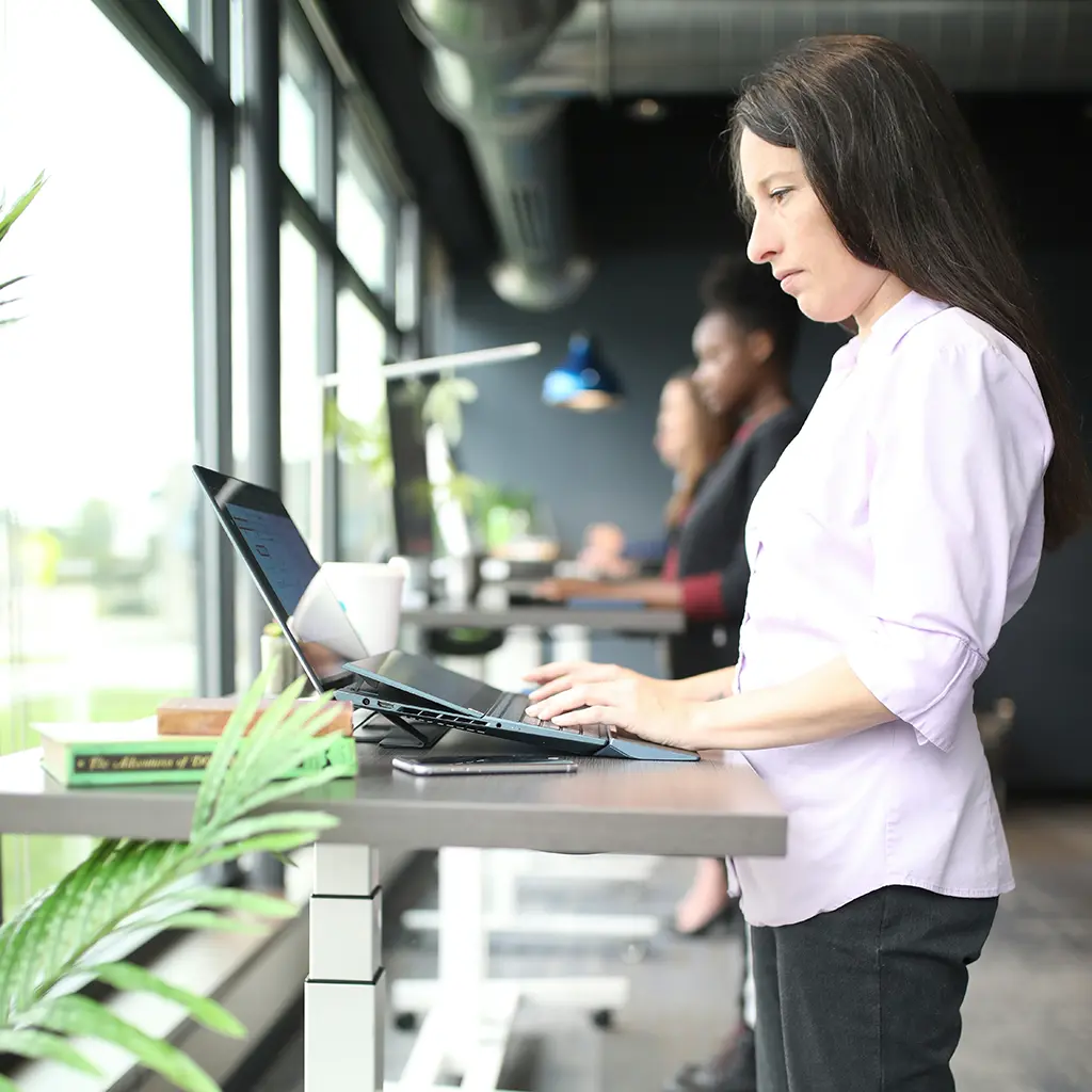 Employee working on a laptop at a height-adjustable standing desk in an open office with coworkers nearby
